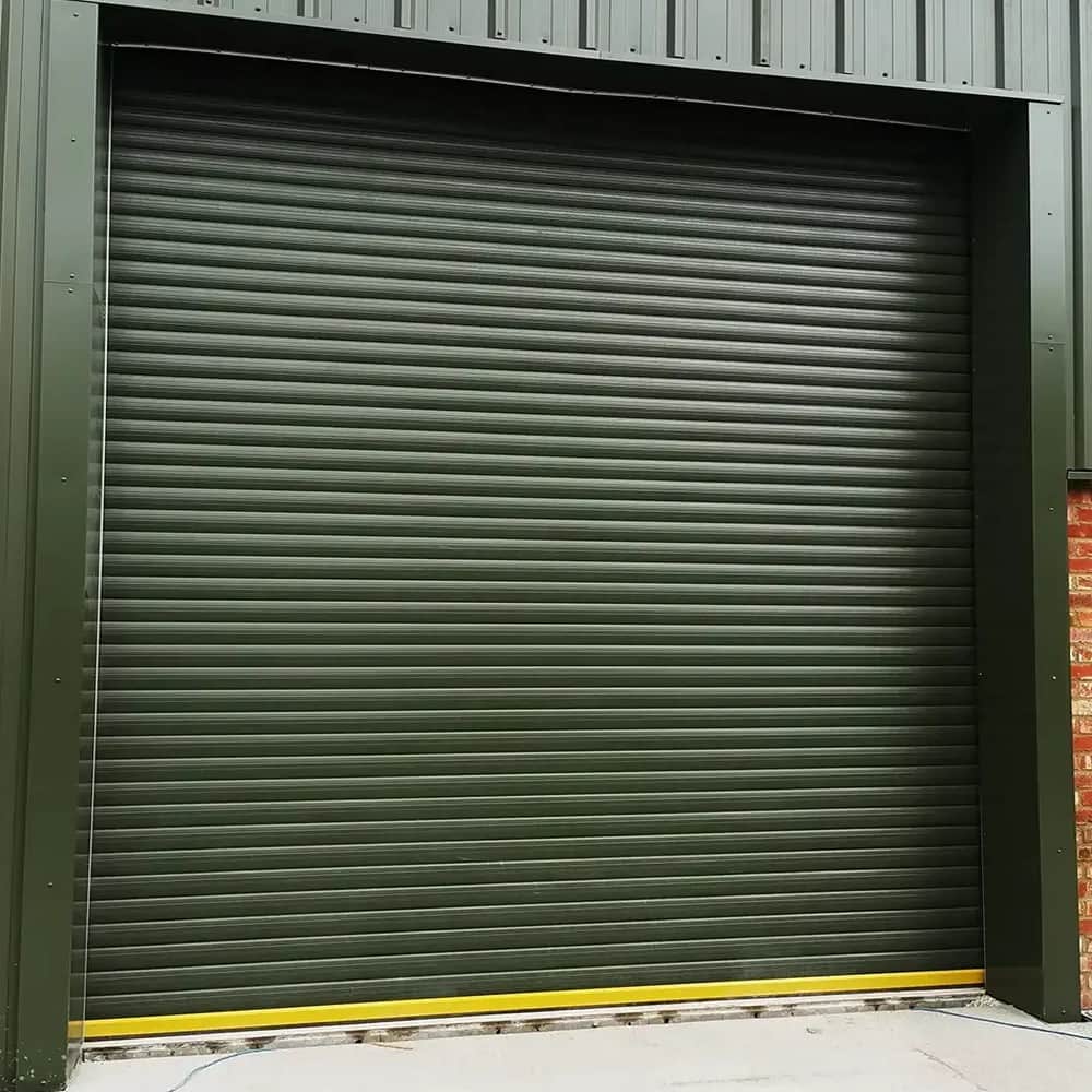 Dark green steel roller shutter closed across a loading opening in a clad industrial unit, with a yellow bottom rail.